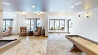 This 700 square foot recreational space in Fort Myers, FL, offers a blend of modern design and diverse entertainment options. The area is characterized by its open layout, featuring a variety of gaming and leisure activities. A prominent shuffleboard table takes center stage, while a professional-grade pool table adds another dimension of entertainment. 

The space boasts a sophisticated design aesthetic with white textured walls, complemented by artistic wall hangings that add visual interest. Lighting is a key feature, with a combination of recessed ceiling lights and statement fixtures, including a striking pendant light over the pool table. Large windows flood the area with natural light and provide views of the surrounding landscape.

Comfortable seating areas are strategically placed throughout, featuring plush armchairs and side tables, ideal for relaxation or spectating. The flooring is a mix of hardwood and patterned carpeting, adding warmth and texture to the space. 

A notable design element is the decorative wall panel with a geometric pattern, which serves as a focal point and adds a touch of modern elegance. The overall ambiance is one of casual luxury, creating an inviting atmosphere for social gatherings and recreational activities.