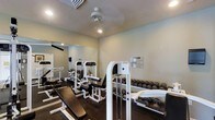 This fitness center is a dedicated single-floor workout facility designed to accommodate a full range of strength and conditioning exercises. The space is outfitted with a selection of white-framed resistance and cable machines, ideal for targeting multiple muscle groups. A well-organized dumbbell rack lines one wall, holding a comprehensive range of free weights for versatile training options. A workout bench is positioned centrally, offering additional functionality for both free weight and bodyweight exercises. Large wall-mounted mirrors enhance the sense of space while allowing users to monitor their form during workouts. The room features recessed lighting paired with a ceiling fan, ensuring the environment remains bright and well-ventilated throughout use. Light-colored walls contribute to a clean, open atmosphere, while the dark flooring provides a strong visual contrast and a stable surface underfoot. A posted workout guide on the wall adds a practical, user-friendly touch. Overall, this fitness center is thoughtfully arranged to maximize usability, offering residents or guests a convenient and fully functional space to maintain their fitness routines.