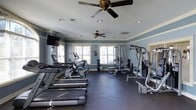 This single-floor fitness center on Donerail Way in Raleigh, NC, offers a well-equipped workout space designed for both cardio and strength training. The room is finished with cool blue-gray walls accented by white crown molding, creating a clean and energizing atmosphere. Recessed lighting, combined with two ceiling fans, ensures the space remains well-lit and comfortable during workouts.

Natural light enters through multiple windows along the far wall, providing views of the surrounding outdoor greenery and adding an open, airy feel to the interior. The dark rubber flooring throughout is both durable and practical, offering cushioning suited for exercise use.

Cardio equipment includes treadmills and stationary bikes positioned toward one side of the room, while a multi-station strength training machine and a weight bench occupy the opposite area, allowing for efficient use of the available floor space. A wall-mounted television is positioned for easy viewing during cardio sessions. A water station is also visible near the windows, adding a convenient amenity for users. Overall, the space is thoughtfully arranged to support a well-rounded fitness routine.