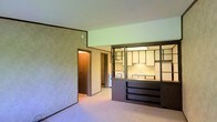 This single-floor residence presents a straightforward and functional layout designed for comfortable everyday living. The main living area is open and airy, featuring light-toned carpeting and textured walls that lend a warm, understated character to the space. A standout architectural detail is the dark-framed built-in shelving unit that serves as a partial room divider, seamlessly connecting the living area to the kitchen while maintaining an open, flowing feel. The kitchen is visible beyond this divider and is equipped with light-colored upper cabinetry, open shelving, and a sink positioned beneath under-cabinet lighting, creating a bright and practical cooking space. The lower portion of the built-in unit provides additional open shelving, offering convenient storage and display space accessible from the living area. A hallway visible from the main room leads to additional areas of the home, suggesting a thoughtfully arranged floor plan. The neutral color palette throughout — from the carpeted floors to the walls and cabinetry — creates a cohesive, calm atmosphere that provides a versatile canvas for a variety of interior styles. Overall, this residence offers a practical and connected single-level living experience.