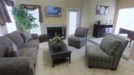This single-floor space centers around a well-appointed living area designed for comfort and functionality. The room features a tile-surround fireplace as a focal point, flanked by windows that allow generous natural light to fill the space. Warm, neutral-toned walls in soft yellow complement the light carpeted flooring, creating an inviting atmosphere throughout.

The living area is generously sized, accommodating a full seating arrangement that includes a sofa, armless accent chairs with striped upholstery, and an additional lounge chair. A dark-finished rectangular coffee table anchors the seating group, topped with a decorative floral arrangement that adds a touch of warmth.

A wall-mounted flat-screen television is positioned above and to the side of the fireplace, while a dark wood media console sits nearby. Decorative touches, including a large wall clock, framed artwork, and a potted plant near the windowed corner, contribute to the cohesive, lived-in aesthetic. A glass-paned door provides additional natural light and suggests access to an exterior area. Overall, the space presents a relaxed yet organized layout suited for everyday living.