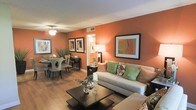 This single-floor residence offers a cohesive and inviting open-plan layout that seamlessly connects the living and dining areas into one harmonious space. The most striking architectural feature is the bold terracotta-orange accent walls, which lend warmth and a distinct character to the interior, complemented by crisp white ceilings and trim throughout.

The living area is anchored by a spacious light-gray sofa adorned with green and floral accent pillows, paired with a dark-finished coffee table that adds contrast. Framed wall art echoes the warm color palette, tying the room's aesthetic together. A ceiling fan provides both function and style overhead.

The dining area sits adjacent to the living space and features a round dining table with upholstered high-back chairs, creating an elegant yet comfortable setting. A decorative plant and framed artwork on the walls enhance the area's refined ambiance.

Rich hardwood-style flooring flows throughout the space, grounding the warm tones and adding a sense of continuity. Soft, layered lighting from ceiling fixtures and table lamps creates a welcoming atmosphere both day and evening. A glimpse of an adjoining hallway or room suggests additional private spaces beyond the main living area.