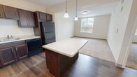 Located at 803 First St in Canonsburg, PA, this one-bedroom, one-bathroom unit offers a well-planned layout across a single floor. The open-concept kitchen serves as the heart of the space, showcasing rich dark wood cabinetry with ample storage, light-toned countertops, and a full suite of black appliances including a range, dishwasher, and refrigerator. A center island with pendant lighting above provides additional prep space and a casual dining option. Durable hardwood-style flooring runs throughout the kitchen and living areas, adding warmth and continuity to the space. The bedroom is a comfortable retreat, finished with plush carpeting and a window that invites natural light and views of the surrounding greenery. A closet with an arched-panel door provides dedicated storage, while a secondary door connects to the rest of the unit. The bathroom features a walk-in shower with a sliding glass enclosure, dark wood vanity cabinetry with a white countertop, and hardwood-style flooring that ties the space together. Neutral tones throughout the unit create a clean, cohesive aesthetic that is both functional and inviting.