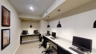 This single-floor business center in Fort Worth is designed to provide a functional and professional environment for work and productivity. The space features a long, continuous white desk surface that runs along the wall, accommodating multiple computer workstations equipped with monitors, keyboards, and mice — offering ample room for guests or tenants to work comfortably side by side. Ergonomic task chairs with green accents and adjustable black frames are positioned at each station, prioritizing comfort during extended use. Two striking black industrial-style pendant lights hang from the ceiling on chain mounts, casting focused light over the workspace and adding a refined, contemporary aesthetic. The neutral color palette — featuring warm beige walls, light-toned flooring, and a crisp white ceiling — creates a calm and distraction-free atmosphere. Overhead HVAC vents are integrated neatly into the ceiling, ensuring a comfortable climate throughout. The overall design strikes a balance between modern style and practical utility, making this business center a well-equipped and inviting space for those requiring a professional setting to handle work, research, or communications.