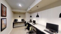 This single-floor business center in Fort Worth is designed to provide a functional and professional environment for work and productivity. The space features a long, continuous white desk surface that runs along the wall, accommodating multiple computer workstations equipped with monitors, keyboards, and mice — offering ample room for guests or tenants to work comfortably side by side. Ergonomic task chairs with green accents and adjustable black frames are positioned at each station, prioritizing comfort during extended use. Two striking black industrial-style pendant lights hang from the ceiling on chain mounts, casting focused light over the workspace and adding a refined, contemporary aesthetic. The neutral color palette — featuring warm beige walls, light-toned flooring, and a crisp white ceiling — creates a calm and distraction-free atmosphere. Overhead HVAC vents are integrated neatly into the ceiling, ensuring a comfortable climate throughout. The overall design strikes a balance between modern style and practical utility, making this business center a well-equipped and inviting space for those requiring a professional setting to handle work, research, or communications.