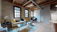 This thoughtfully designed single-floor loft showcases a compelling blend of industrial heritage and contemporary living. The open-concept living and dining area serves as the heart of the space, where original exposed brick walls and heavy wood beam ceilings with visible ductwork create an authentic warehouse aesthetic. Large, black-framed multi-pane windows flood the area with natural light and offer views of the surrounding historic brick architecture, enhancing the urban character of the space. Light hardwood flooring runs throughout the main living areas, grounding the industrial elements with warmth.

The well-appointed kitchen features rich wood cabinetry, a full suite of stainless steel appliances, a peninsula counter, and pendant lighting that echo the exposed-beam ceiling above — making it as functional as it is stylish.

The bedrooms offer a quieter retreat. One bedroom highlights the same exposed brick and large windows found throughout, continuing the loft's signature aesthetic, while another provides a more serene, softly finished environment with neutral walls, plush carpeting, and a built-in closet.

The full bathroom is clean and modern, finished with large-format light tile flooring, a tub-shower combination, and warm wood vanity cabinetry with stone-look countertops. Together, these spaces deliver a cohesive, character-rich living environment.