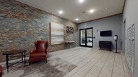 320 Valley presents a well-appointed lobby and common area space designed with both aesthetics and functionality in mind. The entrance lobby immediately establishes a polished, modern identity, anchored by a dramatic stacked stone accent wall in earthy tones of gray, rust, and gold. The building's bold "320 Valley" signage is mounted directly onto this feature wall, creating a strong first impression. Light-toned large-format tile flooring transitions seamlessly into a textured area rug, defining a comfortable lounge seating area furnished with rich cognac leather wingback chairs and a round side table, offering residents a welcoming space to gather.

Recessed lighting throughout the lobby ensures consistent illumination, while a wall-mounted flat-screen television and a sleek cabinet add modern convenience near the double-glass doors leading to the attached parking structure. The lobby also includes a hand sanitizer station and intercom system, reflecting thoughtful attention to resident needs.

Direct access to a generously sized covered parking garage is available through the glass double doors, where concrete structural columns and bright overhead lighting accommodate numerous vehicles with ease. Adjacent to the garage entrance, residents will find a stainless steel elevator and a built-in bank of individual mailboxes, rounding out a comprehensive and efficiently organized ground-floor common area experience.