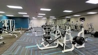 This fitness center is designed to accommodate a wide range of exercise needs within a single, open-plan floor. The space features a patterned carpet in grey and teal tones, providing a clean and modern aesthetic throughout. Overhead, a drop ceiling with evenly spaced recessed panel lighting ensures bright, consistent illumination across the entire area.

The strength training section is prominently featured, with multiple weight benches, barbell racks loaded with plate weights, and a cable/functional trainer machine positioned toward the far wall. A dedicated free weights area offers dumbbells and additional resistance equipment, supporting both beginner and advanced training.

Along the mirrored wall, a row of cardio machines — including treadmills and ellipticals — is neatly arranged, with the mirrors enhancing the sense of depth and space. The mirrored wall also allows users to monitor their form during workouts.

The walls are painted in neutral grey tones with a bold blue accent wall adding a energetic visual contrast. The layout is open and uncluttered, allowing for comfortable movement between equipment zones and making the space feel both functional and welcoming.