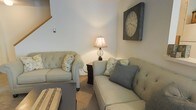 This two-story home offers a comfortable and well-appointed living environment with a charming, classic aesthetic. The living room serves as a central gathering space, featuring plush, tufted upholstered sofas in a soft neutral tone, accented with decorative throw pillows in muted yellows and grays. A rustic, oversized wall clock mounted in a distressed wood frame adds a distinctive focal point to the room, complementing the overall warm and homey atmosphere. Soft carpeting underfoot contributes to the cozy feel of the space, while a table lamp with a classic shade provides warm ambient lighting. A dark end table between the sofas adds a subtle contrast to the light-colored furnishings. The staircase, partially visible in the corner, connects the main living area to the second floor, reinforcing the home's multi-level layout. The walls are finished in a clean, neutral white that enhances the sense of openness and brightness throughout the room. Overall, the space strikes a balance between casual comfort and tasteful décor, making it a welcoming and functional living environment.