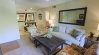 This single-floor residence offers a thoughtfully designed open-concept layout that seamlessly connects the living and dining areas, creating a cohesive and inviting atmosphere. The living room is anchored by a rustic dark wood coffee table paired with a plush white sofa accented with green and neutral throw pillows, complemented by patterned accent chairs that add a touch of personality. A large dark-framed mirror mounted above the sofa enhances the sense of space and light, while recessed lighting and a ceiling fan provide both function and comfort overhead. Soft, neutral-toned carpet flows throughout, contributing to the warm and relaxed ambiance of the home. Framed wall art adds a decorative touch near the dining area, where a dark wood table and stools create an intimate setting for meals. A corridor leading toward what appears to be a kitchen or utility area is visible in the background, hinting at additional functional spaces beyond. Neutral beige walls and subtle architectural details tie the entire space together, making it feel both practical and welcoming. Overall, this home presents a balanced blend of comfort and style within a compact, efficiently arranged single-level floor plan.