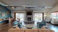 Located at 16102 East Broadway Avenue in Spokane Valley, this single-story community clubhouse offers a well-appointed gathering space designed for both relaxation and socializing. The main lounge area serves as the centerpiece, featuring a striking floor-to-ceiling stacked-stone fireplace with a mounted television above, flanked by large sliding glass doors that flood the room with natural light and open directly to a covered outdoor patio. Polished concrete flooring runs throughout, complemented by warm wood trim accents.

A dedicated dining area adjoins the main lounge, furnished with a solid wood table and chairs, with additional sliding door access to the exterior. The full kitchen is equipped with a gas range, full-size refrigerator, granite-style countertops, and ample wood cabinetry beneath soaring vaulted ceilings accented by bold teal walls and decorative wood art.

The entry foyer features vaulted ceilings, oversized windows offering views of the surrounding neighborhood, and a welcoming seating arrangement of upholstered accent chairs. A secondary lounge area showcases an exposed stacked-stone accent wall, tray ceiling, and flexible seating. An additional common room near the fitness area — visible through interior windows — provides further space for resident use, completing this thoughtfully designed community hub.
