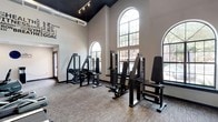 This fitness center is a thoughtfully designed workout facility that combines functional equipment with striking architectural details. The space is defined by soaring ceilings finished in a bold dark tone, exposed ductwork, and track lighting that lends the room an industrial-modern character. Tall, arched windows line the walls, allowing abundant natural light to fill the interior and offering views of the surrounding outdoors.

The facility is organized to accommodate a full range of training needs. A dedicated strength training area features multiple BIOS selectorized resistance machines, including cable stations and a variety of isolation units suited for both upper and lower body workouts. A free weight station offers a tiered dumbbell rack stocked with a wide selection of weights, positioned in front of a large wall mirror adorned with motivational fitness-themed typography — a design detail that reinforces the energetic atmosphere throughout the space.

The cardio zone is equipped with a lineup of TRUE brand treadmills and elliptical machines, each paired with wall-mounted flat-screen televisions for entertainment during workouts. The flooring throughout is durable, textured carpet tile suited for high-traffic athletic use. Together, the equipment variety, generous proportions, and bright, uplifting design make this a comprehensive and inviting fitness destination.