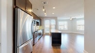 This well-appointed single-floor residence offers a thoughtfully designed open-concept layout that seamlessly connects the kitchen and living areas. The kitchen is a standout feature, showcasing rich dark espresso cabinetry, granite countertops, a large central island with built-in shelving, and a full suite of stainless steel appliances including a refrigerator, range, microwave, and dishwasher. Track lighting overhead highlights the workspace, while wood-look flooring flows throughout the main living areas, adding warmth and continuity to the space. The living area benefits from multiple windows dressed with blinds, inviting generous natural light and featuring a ceiling fan for added comfort. A dedicated hallway leads to the private areas of the home, including a carpeted bedroom with double-door closet access and built-in hanging storage. The full bathroom is finished with large-format tile surrounding the tub and shower, a granite-topped vanity with dark wood cabinetry, and a framed mirror. The in-unit laundry room is a practical highlight, equipped with a stacked washer and dryer, granite folding counter, dark cabinetry, and tile flooring—providing both functionality and style in one convenient space.