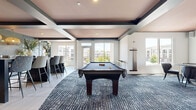 Located at 11240 Woolwich St in Charlotte, NC, this single-floor community amenity space offers a well-appointed recreational and social environment. The open layout is anchored by a full-size billiards table set atop a richly textured, dark-toned area rug, complemented by a shuffleboard table for additional entertainment. A distinctive tray ceiling finished in a warm blush tone, framed by dark geometric beams with recessed lighting, adds architectural character and visual depth to the space.

Natural light pours in through large windows on multiple walls, offering views of the surrounding community and greenery. The flooring transitions between light tile and carpeted areas, delineating zones for gaming and casual lounging. A cozy seating nook features a round table with upholstered chairs, wall art, and a cushioned bench, ideal for small gatherings. A wall-mounted flat-screen TV and a cue rack mounted to the wall enhance the room's functionality.

The entry area features glass doors, tile flooring, and a mounted display screen, while an accent wall with dark wainscoting and decorative wallpaper adds a sophisticated touch. Overall, the space strikes a balance between modern design and comfortable social utility.