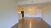 This single-floor unit showcases an open-concept layout that seamlessly connects the kitchen and living areas, creating a cohesive and functional living space. Warm-toned hardwood flooring extends throughout, lending continuity and a sense of warmth to the interior.

The kitchen is positioned to the side and defined by a peninsula counter that serves as both a workspace and a casual dining surface. Warm wood cabinetry lines the upper walls, complemented by black appliances, including a full-size refrigerator. Recessed lighting illuminates the kitchen area, while a pendant light hangs above the peninsula, adding a focused decorative accent.

The open living area is generously proportioned, offering flexibility in furniture arrangement. Neutral white walls and smooth ceilings contribute to a clean, minimalist aesthetic. A tray ceiling detail above the kitchen zone adds subtle architectural interest. Recessed HVAC vents are integrated into the ceiling for a streamlined appearance.

A white entry door is visible at the far end of the living space, completing the layout. Overall, the unit presents a bright, well-maintained interior with a practical and inviting design.