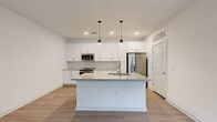 This single-floor, one-bedroom, one-bathroom unit offers a well-designed layout that maximizes its compact footprint. The open-concept kitchen and living area serves as the heart of the unit, featuring light wood-style flooring, recessed lighting, and a central kitchen island with an undermount sink and pendant lighting above. White shaker-style cabinetry extends to the ceiling, complemented by a hexagonal tile backsplash, neutral quartz-style countertops, and a full suite of stainless steel appliances including a range, over-the-range microwave, and French door refrigerator. The living area receives natural light through a large window and front door with transom window. The bedroom is carpeted and bright, with windows providing natural light and ample wall space. The bathroom is well-appointed with a white vanity, framed mirror, wood-look flooring, and a walk-in closet with built-in shelving. A separate laundry area houses a stacked washer and dryer unit. Throughout the space, a consistent palette of white walls, neutral tones, and clean finishes creates a cohesive, modern aesthetic.