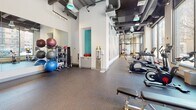 Located at 95 Mathewson St in Providence, Rhode Island, this fitness center offers a thoughtfully designed, single-level workout facility that balances functionality with a modern industrial aesthetic. The space features exposed ductwork and beam ceilings with track lighting overhead, contributing to an open, loft-like atmosphere throughout.

The cardio zone is positioned along a generous bank of floor-to-ceiling windows that overlook the surrounding downtown streetscape, allowing abundant natural light to fill the space while providing views of the Providence cityscape. Treadmills, an elliptical, and a rowing machine are strategically arranged to take full advantage of these views.

A dedicated strength training area houses a comprehensive multi-functional cable and resistance machine, suitable for a wide range of exercises. Nearby, a well-stocked rack holds an assortment of kettlebells and exercise balls, accompanied by a large wall mirror to support proper form. A tiered dumbbell rack is conveniently positioned beneath one of the large windows.

The flooring throughout consists of durable rubberized material suited for high-impact activity. A water station is centrally located for easy access. Glass-paneled partitions add a sleek, contemporary design element, while large-scale wall art enhances the overall ambiance of the space.
