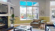 This single-floor community clubhouse offers a thoughtfully designed layout that blends comfort and functionality across several distinct areas. The main lounge is a standout space, featuring soaring ceilings, crown molding, and a two-tone wall treatment of warm yellow-green and cool gray that adds architectural character throughout. A classic fireplace with a decorative mantel and built-in arched niche serves as a focal point, while large floor-to-ceiling windows flood the room with natural light and provide views of the outdoor courtyard and pool area, complete with vibrant shade sails. Warm hardwood flooring runs throughout the main areas, anchored by area rugs that define each seating zone. Adjacent to the lounge, a casual dining area connects seamlessly to a fully equipped kitchen featuring stainless steel appliances, dark granite countertops, white cabinetry, and a breakfast bar with counter-height seating. A secondary gathering room offers a more intimate setting, decorated with ornate carved wood wall panels and a colorful patterned area rug, furnished with small tables and lounge chairs suited for quiet conversation or individual use. Additional seating nooks with natural light from corner windows round out the space, making it a versatile and welcoming environment for a variety of social activities.