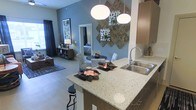 This single-floor residence showcases an open-concept design that integrates the kitchen, dining, and living areas into one cohesive, flowing space. The kitchen is anchored by a generously sized island topped with speckled quartz countertops, offering both prep space and casual seating via bar-height stools. Warm wood-toned cabinetry pairs elegantly with a stainless steel double-basin sink, while two large globe pendant lights hang overhead, providing focused illumination and a contemporary aesthetic touch.

The dining area sits just beyond the island, featuring a round table with patterned upholstered chairs beneath a striking wall arrangement of textured, geometric art panels. Cool blue-grey walls create a calm, modern backdrop throughout the space, complementing the light-toned flooring that runs continuously underfoot, enhancing the sense of openness.

The living area extends toward the far end, where natural light filters in through a large window dressed with dark curtains. A full-length mirror near the dining space adds depth and brightness. Overall, the space presents a well-appointed, stylish atmosphere with a balanced mix of modern fixtures and warm design elements.