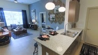 This single-floor residence showcases an open-concept design that integrates the kitchen, dining, and living areas into one cohesive, flowing space. The kitchen is anchored by a generously sized island topped with speckled quartz countertops, offering both prep space and casual seating via bar-height stools. Warm wood-toned cabinetry pairs elegantly with a stainless steel double-basin sink, while two large globe pendant lights hang overhead, providing focused illumination and a contemporary aesthetic touch.

The dining area sits just beyond the island, featuring a round table with patterned upholstered chairs beneath a striking wall arrangement of textured, geometric art panels. Cool blue-grey walls create a calm, modern backdrop throughout the space, complementing the light-toned flooring that runs continuously underfoot, enhancing the sense of openness.

The living area extends toward the far end, where natural light filters in through a large window dressed with dark curtains. A full-length mirror near the dining space adds depth and brightness. Overall, the space presents a well-appointed, stylish atmosphere with a balanced mix of modern fixtures and warm design elements.
