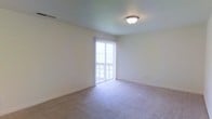 This single-floor residential space is designed with simplicity and functionality in mind. The interior is finished with neutral, light-toned walls that create a clean and airy atmosphere throughout. The living area features wall-to-wall carpeting in a warm beige tone, providing a comfortable and welcoming feel underfoot. A sliding glass door, dressed with vertical blinds, serves as a central architectural feature of the main room, allowing an abundance of natural light to fill the space while providing direct access to an outdoor balcony. The balcony view hints at a pleasant outdoor setting, adding a sense of openness to the interior. Ceiling-mounted lighting provides consistent illumination across the room, complementing the natural light that streams in. The walls are smooth and well-maintained, framed by clean white baseboards that run throughout. The overall ambiance of the space is calm, bright, and neutral — well-suited for a variety of furnishing styles. The straightforward, open layout of the main living area offers flexibility for arrangement, making it a practical and inviting environment for everyday living.