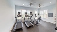 This fitness center is a thoughtfully designed, single-floor facility built to support a comprehensive workout experience. The space is finished with durable rubber flooring throughout, soft blue-gray walls, and recessed lighting that creates a clean, energizing atmosphere. Large windows fitted with white blinds line the cardio area, allowing ample natural light to filter in and offering views of the surrounding outdoor greenery.

The cardio zone is well-appointed with two treadmills, an elliptical cross-trainer, and a recumbent bike, each equipped with individual display screens for entertainment and performance tracking. Ceiling fans overhead ensure comfortable airflow during workouts.

A dedicated strength training area features a multi-functional cable machine paired with an adjustable weight bench and a full-length mirror, ideal for form monitoring during resistance exercises. A rack of dumbbells provides additional free weight options for a variety of routines.

Functional training accessories include a collection of medicine balls displayed on a tiered rack, as well as balance and stability equipment such as BOSU trainers and exercise balls. A wall-mounted flat-screen television adds entertainment value to the strength training zone. Posted fitness center rules and regulations reflect a commitment to a safe and organized environment for all users.