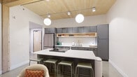 This thoughtfully designed one-bedroom, one-bathroom residence in Sioux Falls, SD, blends modern aesthetics with warm architectural details throughout its efficiently organized single-floor layout. The open-concept living and kitchen area serves as the heart of the home, anchored by a striking natural wood plank ceiling that adds warmth and texture. Polished concrete floors extend throughout the main living spaces, complementing the contemporary palette. The kitchen is a standout feature, boasting sleek charcoal cabinetry, open wood shelving, stainless steel appliances, and a large central island with a white quartz countertop and bar seating — finished with globe pendant lighting for a stylish, modern touch. The living area flows seamlessly toward floor-to-ceiling sliding glass doors that open onto a private balcony, filling the space with natural light and offering views of the surrounding building courtyard. The bedroom is a calm, neutral retreat with soft carpeting, exposed wood ceiling accents, and a built-in closet. The bathroom features hexagonal tile flooring, a white quartz vanity, and a round mirror. A dedicated laundry room with a full-size washer and dryer and built-in bench seating adds everyday convenience to this well-appointed residence.