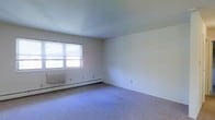 This single-floor space presents a straightforward and functional layout. The room shown is generously proportioned, featuring wall-to-wall carpeting in a neutral tone that provides a soft, comfortable foundation underfoot. The walls are finished in a clean, light off-white, contributing to a bright and open atmosphere. A window along one wall allows natural light to filter into the space, enhancing the airy feel of the room. Baseboard heating units run along the perimeter of the walls, offering an efficient and unobtrusive heating solution. Electrical outlets are conveniently positioned at various points throughout the room, providing practical functionality. The standard ceiling height and simple, clean lines give the space a modest yet versatile character, making it well-suited for a variety of uses. The overall ambiance is understated and neutral, offering a blank canvas that can be easily personalized and arranged to suit individual preferences. The absence of clutter and the minimalist finishes allow the structural elements of the room to speak for themselves, emphasizing practicality and livability.