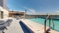 Located in the heart of downtown Madison, Wisconsin, this rooftop terrace at 109 E Wilson Street offers an expansive outdoor retreat with stunning panoramic views of Lake Monona. The centerpiece of the space is a sleek, tiled swimming pool, designed for leisure and surrounded by generous deck space finished in large-format stone pavers. Rows of sun loungers are arranged throughout the terrace, providing ample space for relaxation in the open air.

A glass balustrade perimeter railing preserves the unobstructed lakefront views while maintaining an open, airy feel across the entire rooftop level. Adjacent to the pool area, a dedicated lounge zone features modern outdoor sofas, armchairs, and a wall-mounted flat-screen television, creating a comfortable setting for both socializing and unwinding. Large sliding glass doors connect this lounge area seamlessly to the interior of the building.

Additional amenities include high-top bistro tables and chairs ideal for outdoor dining, a built-in bar counter finished in complementary tile, and multiple lighting fixtures positioned throughout for evening use. The architectural aesthetic is clean and contemporary, with paneled wall cladding in neutral tones that complement the open sky above. This rooftop destination combines resort-style amenities with exceptional urban lakeside views.