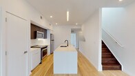 This contemporary two-floor residence showcases a clean, modern design throughout, with light hardwood flooring and crisp white walls creating a bright, cohesive aesthetic across both levels.

The main floor centers on a well-appointed open-plan kitchen, where a generous elongated island with an undermount sink and matte black faucet serves as the focal point. Dark espresso upper cabinetry contrasts with white lower cabinets, while stainless steel appliances — including a range, microwave, and refrigerator — complete the functional layout. A linear pendant light overhead adds a refined touch. The kitchen flows seamlessly toward a staircase with rich dark wood treads, leading to the upper level.

Upstairs, multiple rooms feature expansive floor-to-ceiling windows that frame urban city views, flooding the spaces with natural light. A wide hallway connects the rooms with clean sightlines and continued hardwood flooring. A conveniently tucked in-unit stacked washer and dryer provides added practicality.

Two full bathrooms are finished with large-format neutral stone-look tiles on both the floors and walls, each equipped with a soaking tub and shower combination, matte black fixtures, and a floating vanity — delivering a spa-like atmosphere. The overall design emphasizes minimalism, functionality, and modern urban living.