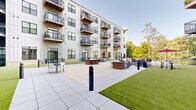 Located at 309 W Front St in the heart of Traverse City, Michigan, this contemporary multi-story building showcases a striking modern architectural design with light-toned panel siding, large dark-framed windows, and wood-accented balconies equipped with sleek metal railings. Spanning two floors, the structure wraps around a generously sized central courtyard that serves as a premier outdoor amenity space.

The courtyard is thoughtfully designed for both relaxation and social gatherings. A central fire pit surrounded by Adirondack-style chairs creates an inviting focal point, while a brick-built outdoor grilling station offers functional space for outdoor cooking and dining. Seating areas with tables and chairs are distributed throughout the paved terrace, complemented by stretches of well-maintained artificial turf that add a vibrant, green contrast to the light concrete pavers.

Perimeter walkways with black iron railings overlook a lush natural tree canopy, providing residents with serene, verdant views and a strong connection to the surrounding landscape. The building's balconies — visible on multiple levels — extend the indoor-outdoor living experience, each offering elevated views of the courtyard and neighboring greenery. The overall ambiance is one of polished urban living harmoniously integrated with nature, ideally positioned in the vibrant downtown Traverse City corridor.