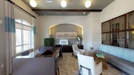 This single-floor lobby offers a polished and inviting atmosphere designed to make a strong first impression. At its center, a dark espresso-toned reception desk anchors the space, providing a clear point of welcome for visitors. The walls are finished in a soft neutral tone, complemented by rich dark wood accents including a prominent ceiling beam that adds architectural character and warmth.

A standout feature is the arched wall niche, backlit to highlight a framed architectural print and a floral arrangement, creating a sophisticated focal point. Dark wood cabinetry flanks this niche, offering functional storage while maintaining the space's refined aesthetic. Globe pendant lights hang from the ceiling, casting a warm, even glow throughout.

The seating area includes patterned upholstered chairs, an ornate accent table, and additional cushioned seating, arranged to encourage comfortable conversation. Natural light filters in through windows dressed with light curtains, brightening the stone-tiled flooring. Decorative greenery adds a fresh, organic touch, contributing to the lobby's overall polished and professional ambiance.