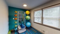 Located at 7025 Bronner Cir in Louisville, Kentucky, this single-floor fitness room is purpose-built to support a well-rounded exercise experience. The space is defined by its vibrant, multi-toned blue and grey carpet tile flooring, which adds both visual energy and comfort underfoot. A bold teal accent wall anchors the main equipment area, creating a motivating and dynamic atmosphere.

The room is outfitted with a Smart-brand multi-function equipment rack that organizes an array of fitness tools, including resistance bands, medicine balls in varying weights, stability balls in multiple sizes, and rolled exercise mats — all neatly stored and easily accessible. A full-length wall mirror with a dark wood frame is mounted alongside a wooden bench, providing a functional area for stretching or changing footwear.

Natural light filters into the space through a double-panel window fitted with horizontal blinds, offering a view of the surrounding greenery outside. Recessed and flush-mounted ceiling lights ensure consistent illumination throughout. The room is accessed through a set of glass-panel double doors, contributing to an open and welcoming entry. The overall design balances practicality with an energizing aesthetic, making this fitness room a well-equipped and inviting space for exercise and wellness activities.