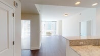This contemporary apartment showcases an open-concept design that maximizes space and light. The main living area boasts rich hardwood flooring that extends throughout, creating a seamless flow between rooms. Large windows flood the space with natural light and offer expansive city views, enhancing the urban living experience.

The kitchen is a highlight, featuring sleek dark cabinetry contrasted with stainless steel appliances and a light-colored granite countertop. A breakfast bar provides additional seating and separates the kitchen from the living area while maintaining the open feel.

The bedroom continues the modern aesthetic with ample natural light and built-in climate control. The bathrooms are tastefully appointed with modern fixtures, including a vanity with undermount sink, large mirror, and a glass-enclosed shower in one, while the other offers a shower/tub combination with accessibility features.

Throughout the apartment, recessed lighting and clean white walls contribute to the bright, airy atmosphere. The layout is efficient, with clever storage solutions and a dedicated laundry area. This apartment embodies contemporary urban living, balancing style with functionality in a compact yet comfortable space.