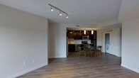 This single-floor residence showcases an open-concept layout that connects the living, dining, and kitchen areas into one cohesive and functional space. The flooring throughout is a warm-toned hardwood-style material that adds continuity and a clean aesthetic across the entire unit.

The kitchen is defined by rich, dark cabinetry that extends to the ceiling, providing generous storage while creating a bold contrast against the lighter walls and countertops. Stainless steel appliances, including a refrigerator, microwave, and range, lend a modern, polished look. Pendant lighting hangs above the kitchen area, offering focused illumination that complements the recessed ceiling lights within the kitchen's slightly raised ceiling detail.

The dining area sits just outside the kitchen, accommodating a mid-sized table and chairs, making it well-suited for everyday dining or casual entertaining. Adjacent to this zone, a stacked washer and dryer unit is conveniently tucked along the wall, adding in-unit laundry functionality.

The living area is open and bright, with track lighting on the ceiling providing adjustable illumination. Neutral gray walls and clean white baseboards maintain a simple, modern aesthetic throughout the space.