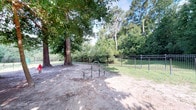 Located at 8439 Dorchester Rd in North Charleston, South Carolina, this single-level outdoor space offers a well-appointed dog park area designed for both pets and their owners. The space is framed by a sturdy black wrought-iron perimeter fence, providing a secure and defined boundary for use. A pair of stainless steel agility hoops serves as an engaging amenity for dogs, positioned on a sandy, natural ground surface that is easy on paws. The area benefits from generous shade provided by large, mature trees — including a prominent tree near the entrance — whose canopies filter sunlight throughout the day, creating a comfortable environment even in warm South Carolina weather. The lush, dense woodland visible beyond the fence adds a serene, natural backdrop, enhancing the sense of seclusion and tranquility. A well-maintained grassy area borders the sandy zone, contributing to a clean and inviting atmosphere. The combination of natural tree coverage, secure fencing, and dedicated pet amenities makes this outdoor space a thoughtfully designed destination for recreation and outdoor enjoyment.