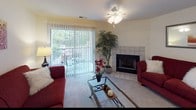 This thoughtfully designed single-floor model home offers a comfortable and functional layout across several well-defined living spaces. The living room serves as the heart of the home, anchored by a tile-surround fireplace and plush carpeting. A ceiling fan with light fixture overhead complements the natural light streaming in through a large sliding glass door fitted with vertical blinds, which opens to an outdoor view. The adjacent dining area features carpet flooring, an elegant chandelier, and a pass-through opening to the kitchen, creating a seamless flow for everyday living and entertaining. The kitchen is equipped with warm wood cabinetry, white countertops, a range with cooktop, and a black refrigerator, all set on easy-care wood-look flooring. Two bedrooms provide restful retreats, each with soft carpet, ample natural light from windows with blinds and curtains, and room for full-sized furnishings. The full bathroom impresses with a wide dual-sink vanity topped in white, rich wood cabinetry below, a large mirror with Hollywood-style lighting, and a tub-shower combination. A dedicated linen closet with louvered doors and built-in wire shelving adds practical storage near the bathroom, rounding out this well-organized and inviting model home.