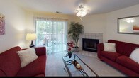 This thoughtfully designed single-floor model home offers a comfortable and functional layout across several well-defined living spaces. The living room serves as the heart of the home, anchored by a tile-surround fireplace and plush carpeting. A ceiling fan with light fixture overhead complements the natural light streaming in through a large sliding glass door fitted with vertical blinds, which opens to an outdoor view. The adjacent dining area features carpet flooring, an elegant chandelier, and a pass-through opening to the kitchen, creating a seamless flow for everyday living and entertaining. The kitchen is equipped with warm wood cabinetry, white countertops, a range with cooktop, and a black refrigerator, all set on easy-care wood-look flooring. Two bedrooms provide restful retreats, each with soft carpet, ample natural light from windows with blinds and curtains, and room for full-sized furnishings. The full bathroom impresses with a wide dual-sink vanity topped in white, rich wood cabinetry below, a large mirror with Hollywood-style lighting, and a tub-shower combination. A dedicated linen closet with louvered doors and built-in wire shelving adds practical storage near the bathroom, rounding out this well-organized and inviting model home.