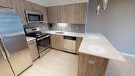 This single-floor unit offers a well-designed layout with modern finishes that create a cohesive, contemporary aesthetic. The kitchen serves as a functional centerpiece, featuring flat-front cabinetry in a warm wood-grain finish, white quartz countertops, a gray subway tile backsplash, and a full suite of stainless steel appliances including a French door refrigerator, electric range, built-in microwave, and dishwasher. A peninsula counter provides additional workspace and separation from the living area. The open living and dining space is finished with light wood-look flooring and neutral warm-gray walls, with track lighting overhead adding a modern touch. A large floor-to-ceiling window allows ample natural light and offers outdoor views of surrounding greenery. The bedroom continues the neutral palette with wall-to-wall carpet, taupe walls, and its own access to natural light through a sizable window. The bathroom is finished in a clean, understated style with a full soaking tub and shower combination, beige tile surround, and brushed nickel fixtures. Throughout the unit, white baseboards and consistent flooring choices contribute to a unified, move-in-ready appearance.