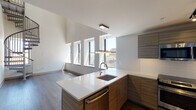 This contemporary two-floor residence showcases a thoughtfully designed open layout that balances modern aesthetics with functional living. The main level centers around a well-appointed kitchen featuring sleek, wood-grain cabinetry, white quartz countertops, a kitchen island with an undermount sink, and a full suite of stainless steel appliances including a range, microwave, dishwasher, and refrigerator. White subway tile backsplash and track lighting complete the polished look. The main living area is defined by rich hardwood flooring and soaring double-height ceilings, with a wall of large windows that flood the space with natural light and frame impressive urban city views. A sculptural metal spiral staircase serves as both a functional and architectural centerpiece, connecting the main level to the upper floor. Upstairs, the secondary level offers carpeted bedroom spaces with generous closet storage and high ceilings that continue the sense of volume throughout. A full bathroom on this level features large-format tile flooring, a frameless glass-enclosed walk-in shower with white subway tile surround, and contemporary wood-tone vanity cabinetry. The overall ambiance is clean, modern, and light-filled, with refined finishes carried consistently throughout both floors.