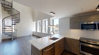 This contemporary two-floor residence showcases a thoughtfully designed open layout that balances modern aesthetics with functional living. The main level centers around a well-appointed kitchen featuring sleek, wood-grain cabinetry, white quartz countertops, a kitchen island with an undermount sink, and a full suite of stainless steel appliances including a range, microwave, dishwasher, and refrigerator. White subway tile backsplash and track lighting complete the polished look. The main living area is defined by rich hardwood flooring and soaring double-height ceilings, with a wall of large windows that flood the space with natural light and frame impressive urban city views. A sculptural metal spiral staircase serves as both a functional and architectural centerpiece, connecting the main level to the upper floor. Upstairs, the secondary level offers carpeted bedroom spaces with generous closet storage and high ceilings that continue the sense of volume throughout. A full bathroom on this level features large-format tile flooring, a frameless glass-enclosed walk-in shower with white subway tile surround, and contemporary wood-tone vanity cabinetry. The overall ambiance is clean, modern, and light-filled, with refined finishes carried consistently throughout both floors.