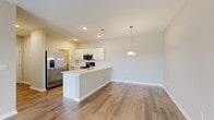 This single-floor residence offers a clean, modern design with a thoughtful layout that balances open living spaces and private retreats. The heart of the home is an open-concept kitchen and living area, where wood-look flooring flows seamlessly underfoot. The kitchen is equipped with stainless steel appliances — including a French door refrigerator with ice maker, a range, and a microwave — complemented by crisp white cabinetry and a spacious center island with an integrated sink and light countertops. A pendant light fixture adds a polished finishing touch above the island. The adjoining living area is bright and airy, featuring a ceiling fan, large windows with horizontal blinds, and a glass-panel door that opens to the outdoors, inviting natural light and views of the surrounding landscape. The bedrooms are carpeted for comfort and include windows that bring in additional natural light. A dedicated laundry area houses a top-load washer alongside built-in wire shelving for organized storage. The bathroom features a full walk-in shower with large white subway tile, a glass sliding door, and chrome fixtures, presenting a clean and contemporary aesthetic. Throughout, neutral wall tones and consistent trim work create a cohesive and inviting atmosphere.