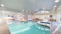 This single-level indoor aquatic facility is designed to offer a refined and relaxing environment for swimmers and spa enthusiasts alike. The centerpiece of the space is a generously sized indoor swimming pool with crystal-clear turquoise water, illuminated by underwater lighting that enhances the serene atmosphere. Adjacent to the main pool, an integrated circular hot tub provides a dedicated space for hydrotherapy and relaxation.

The architecture is defined by striking vaulted ceilings that rise dramatically overhead, lending an open and airy quality to the interior. A continuous band of large, floor-to-ceiling windows and transom windows wraps around a key section of the facility, flooding the space with abundant natural light and offering views of the surrounding wooded landscape. French-style glass doors provide direct access to an outdoor patio area beyond.

A dedicated lounge zone features contemporary modular seating with teal accent cushions, positioned to take full advantage of the natural light and outdoor views. The flooring throughout the lounge and poolside areas consists of polished stamped concrete, complemented by warm stone tile accents along the pool surround and lower walls. Recessed ceiling lighting supplements natural illumination throughout the facility, ensuring a well-lit and inviting environment year-round.