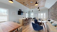 Located at 9 Covered Bridge Rd in Newtown, CT, this single-floor community clubhouse blends rustic charm with contemporary design. The main gathering area is an inviting, open-concept space anchored by a dramatic full-height stacked stone accent wall that adds warmth and texture. Light hardwood flooring runs throughout, complementing soft blue-gray walls and an abundance of natural light streaming in through multiple windows and glass-paneled exterior doors.

The bar area is a standout feature, boasting a wood-paneled peninsula with granite countertops, white shaker cabinetry, a subway tile backsplash, and a stainless steel sink. A wall-mounted TV, open shelving, and a compact refrigerator complete this social hub. Overhead, vaulted wood-planked ceilings with black pendant lights add architectural character and a welcoming ambiance.

The lounge zone offers comfortable seating, including a tufted sofa, plush navy accent chairs, and round wood tables — ideal for casual gatherings or quiet relaxation. A separate seating nook near the entry provides additional intimate space with dark leather chairs and ample natural light.

Adjoining the main lounge is a well-equipped fitness center, visible through a partition wall, offering a range of cardio and strength-training equipment. Together, these spaces create a well-rounded, amenity-rich environment designed for community engagement and everyday comfort.