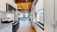 Located at 800 Broadway Blvd in the heart of Kansas City, MO, this urban loft blends historic industrial character with modern finishes. The single-floor layout includes a combined kitchen and living area along with one full bathroom, making efficient use of a compact but thoughtfully designed footprint.

The open living space is defined by striking exposed wood beam ceilings and warm hardwood floors that run throughout. Multiple large windows fitted with roller shades line the walls, flooding the interior with natural light and offering views of the surrounding downtown Kansas City streetscape. A wall-mounted mini-split unit provides climate control, while a ceiling fan adds additional airflow to the airy space. Slate-blue accent walls provide depth and contrast to the bright white trim and walls.

The galley-style kitchen is well-appointed with white cabinetry, granite countertops, a subway tile backsplash, and a full suite of stainless steel appliances including a range, microwave, refrigerator, and dishwasher. A sliding barn door and industrial-style double doors add to the loft's character.

The bathroom continues the clean aesthetic with white cabinetry, a white vanity countertop, a walk-in tiled shower with a sliding glass door, and hardwood flooring. The overall ambiance is a seamless marriage of historic urban architecture and contemporary urban living.