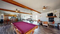 This single-story clubhouse offers a generously proportioned, open-plan layout designed for social gatherings and recreation. Exposed wood ceiling beams run the length of the main area, adding warmth and architectural character beneath vaulted ceilings fitted with multiple ceiling fans. Dark hardwood-style flooring flows throughout, unifying each zone seamlessly.

The main gathering space is anchored by a full-sized pool table with a bold magenta felt surface, complemented by a ping pong table nearby, making it a central hub for entertainment. Adjacent to the game area, a comfortable lounge zone features a dark leather sofa, organic-shaped coffee table, and large windows that admit ample natural light and views of the surrounding community.

A stone-clad fireplace wall dominates one corner, topped with a flat-screen television and flanked by mid-century-style chairs. Glass doors from this seating area open directly to an outdoor balcony. A dining area with a raised table and bar-style seating sits near a glass entry door, surrounded by wide windows overlooking the exterior grounds.

The kitchen is well-equipped with warm oak cabinetry, granite countertops, stainless steel appliances including a refrigerator, range, microwave, and dishwasher, and a curved breakfast bar with bar stool seating. The overall ambiance is casual, welcoming, and functional.