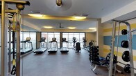 This fitness center offers a thoughtfully designed workout space with a clean, modern aesthetic. The room features large windows along one wall, flooding the space with natural light and providing views of the surrounding rooftop and cityscape beyond. Flush circular light fixtures with warm yellow accents are recessed into a dropped ceiling detail, complementing the room's bright and energizing atmosphere. A ceiling fan aids in air circulation throughout the space.

The exercise floor is finished with durable grey fitness flooring, providing a practical and comfortable surface for workouts. A row of treadmills lines the windowed wall, each equipped with individual digital display screens. Alongside these, elliptical trainers offer additional cardiovascular options. On the opposite side of the room, a well-stocked dumbbell rack holds a full range of free weights, accompanied by a weight bench for strength training. A full-length mirror is positioned along one wall, enhancing the sense of space. The overall design combines functionality with a clean, contemporary look, incorporating yellow and grey tones that contribute to a motivating and organized workout environment.