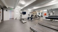 Located at 8000 E Girard Ave in Denver, Colorado, this fitness facility offers a comprehensive workout environment across a single floor. The space is divided into a primary exercise room, a dedicated multi-purpose yoga and activity room, and a connecting hallway.

The main exercise room features durable dark rubber flooring and is outfitted with a robust selection of cardio equipment, including multiple True-brand treadmills, elliptical trainers, a recumbent bike, and additional strength and conditioning machines. Wall-mounted sconces provide even ambient lighting alongside flush ceiling fixtures, while full-wall mirrors along one side expand the sense of space and allow users to monitor their form. A wall-mounted TV adds convenience for those on cardio equipment.

The adjoining yoga and multi-purpose room is bright and open, featuring smooth flooring ideal for yoga, stretching, or group exercise. This room is equipped with a wall-mounted Swedish ladder, a collection of exercise balls in various sizes, and a ping-pong table, making it highly versatile. Floor-to-ceiling glass partition walls connect the two main areas, promoting an open, airy feel throughout the facility.

The design throughout is clean and modern, with neutral gray tones, structured ceiling beams, and a functional layout that maximizes usable floor space for a variety of fitness activities.