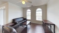 This single-floor residence offers a thoughtfully laid-out living environment across several distinct spaces. The living room is a standout area, highlighted by a pair of arched transom windows that flood the room with natural light, crown molding, durable hardwood-style flooring, and a ceiling fan overhead. A leather sofa and armchair complete the inviting atmosphere.

The kitchen is well-equipped with white cabinetry featuring brass hardware, a double-basin stainless steel sink, laminate countertops, a full suite of white appliances including a range, microwave, and refrigerator, and track lighting above. Hardwood-style flooring continues throughout this space.

Adjacent to the kitchen, the dining area accommodates a round table with seating for four, brightened by vertical blinds along a wide window wall and track lighting above.

The bedrooms are carpeted and equipped with ceiling fans. One bedroom features a distinctive arched window with views to the exterior, adding architectural character. Each room offers comfortable proportions suitable for study and rest.

The bathroom includes a vanity with a white cabinet base, a large mirror, a window for natural ventilation, and a separate tub-shower combination with recessed lighting. The space maintains a clean, neutral palette throughout, creating a cohesive and functional home environment.