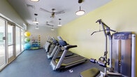 This fitness center offers a comprehensive workout environment designed to support a wide range of exercise routines. The space is defined by bold yellow accent walls paired with crisp white ceilings, creating an energizing atmosphere conducive to physical activity. Durable dark rubber flooring covers the entire area, providing both safety and resilience underfoot.

The cardio section is well-stocked with multiple treadmills and elliptical machines, each equipped with individual digital displays to support performance tracking. A stationary bike rounds out the aerobic options. For strength training, the space includes several selectorized cable and weight machines, a multi-functional cable station, and an adjustable workout bench, offering a solid range of resistance training choices.

A dedicated functional fitness rack along one wall holds an organized assortment of medicine balls, resistance bands, and foam rollers, making it easy to incorporate dynamic and recovery-focused exercises into any routine. Overhead, multiple ceiling fans and pendant lights work in tandem to maintain a comfortable environment and ensure consistent, well-distributed illumination throughout the space. The overall layout is open and efficiently arranged, allowing users to move freely between equipment stations.